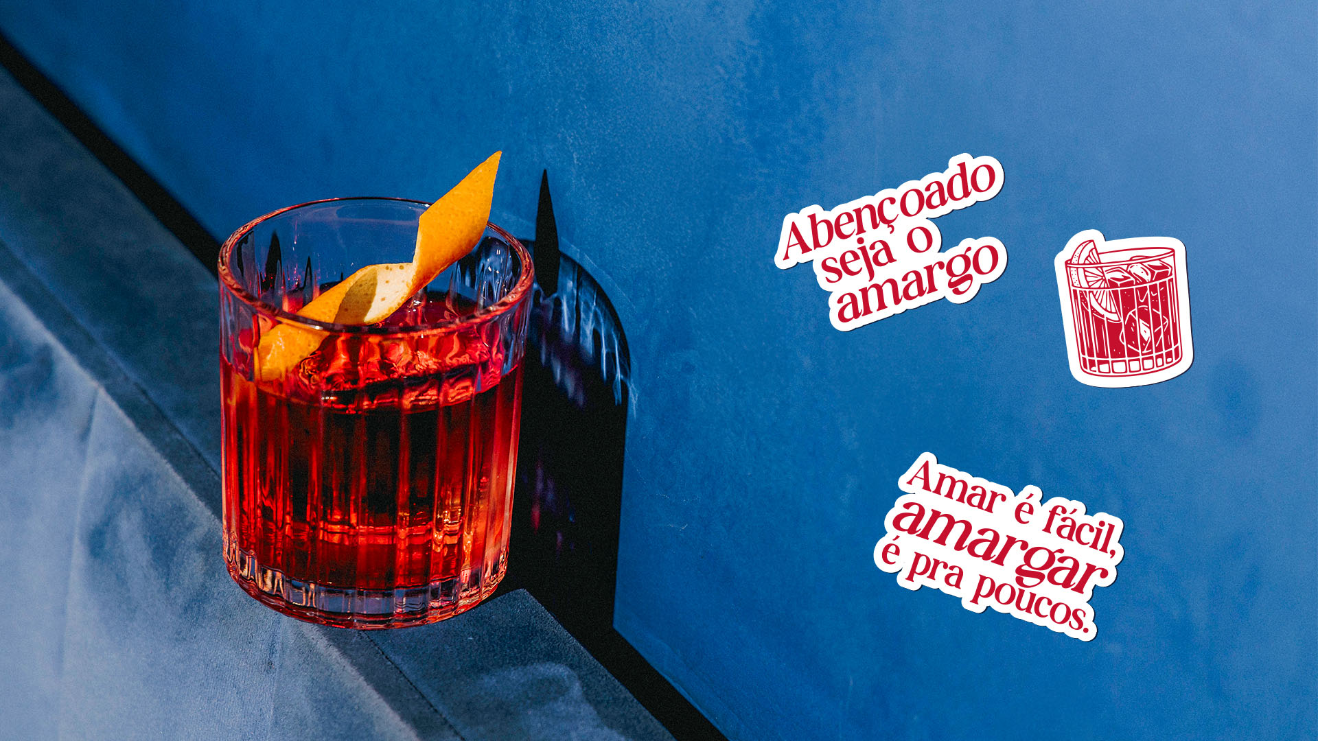Negroni em copo canelado sobre superfície azul, com adesivos Abençoado seja o amargo e Amar é fácil, amargar é pra poucos